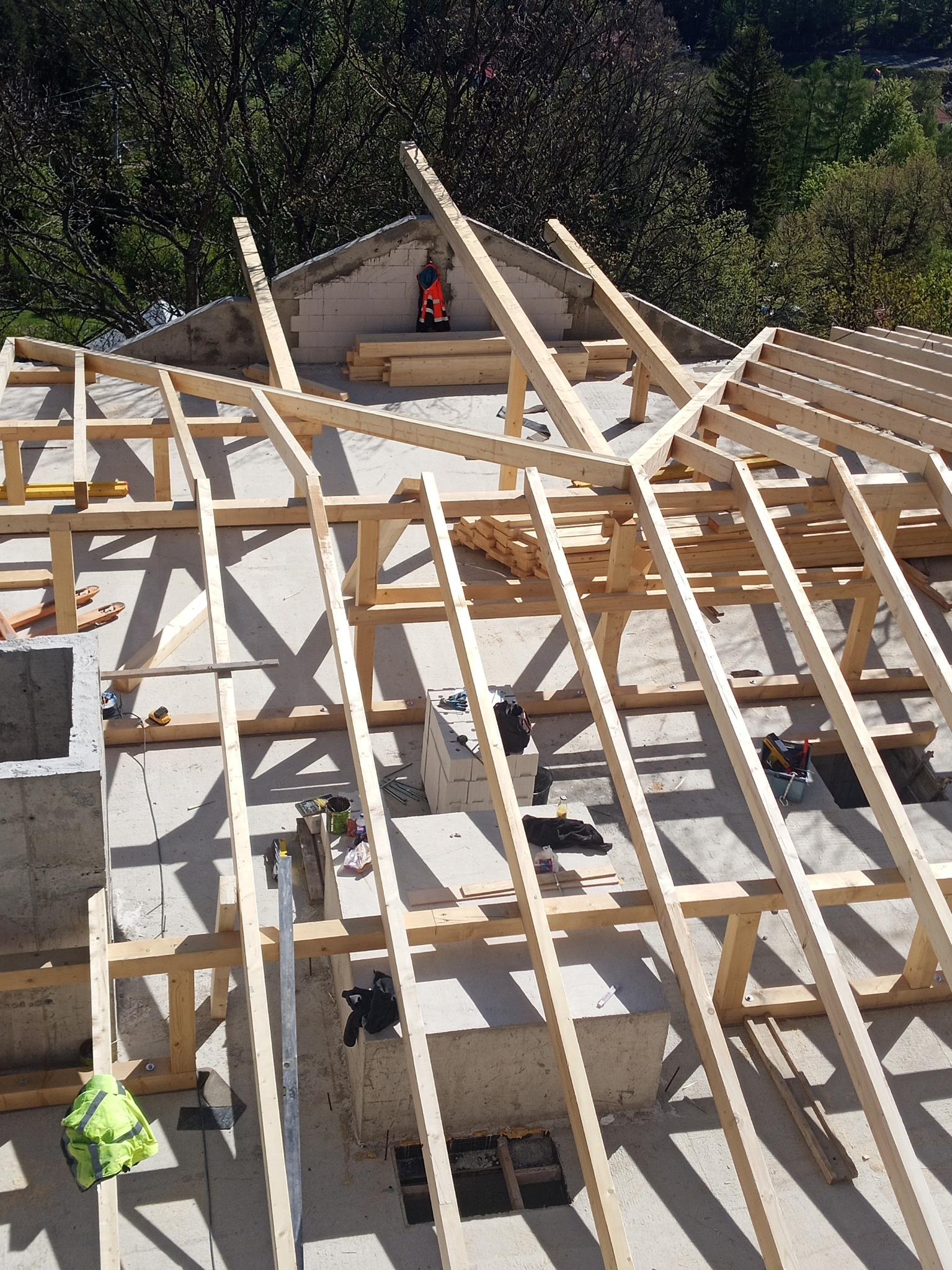 Wysoki kąt: drewniana więźba dachowa w trakcie montażu na betonowym budynku, widoczne narzędzia i elementy konstrukcyjne, drzewa w tle.