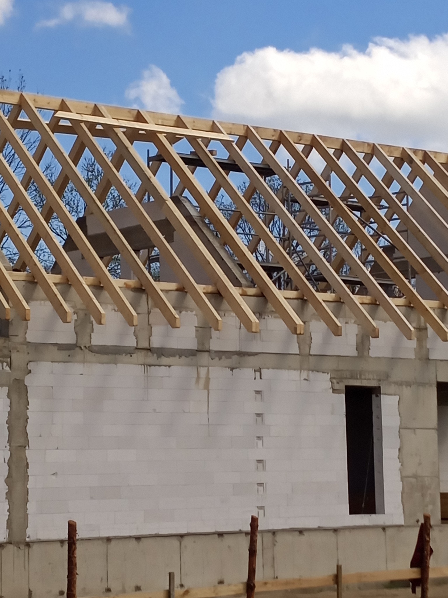 Wznoszenie więźby dachowej na budynku z białych bloczków, widoczne niebo i drzewa w tle. Konstrukcja w trakcie budowy, drewniane elementy więźby.