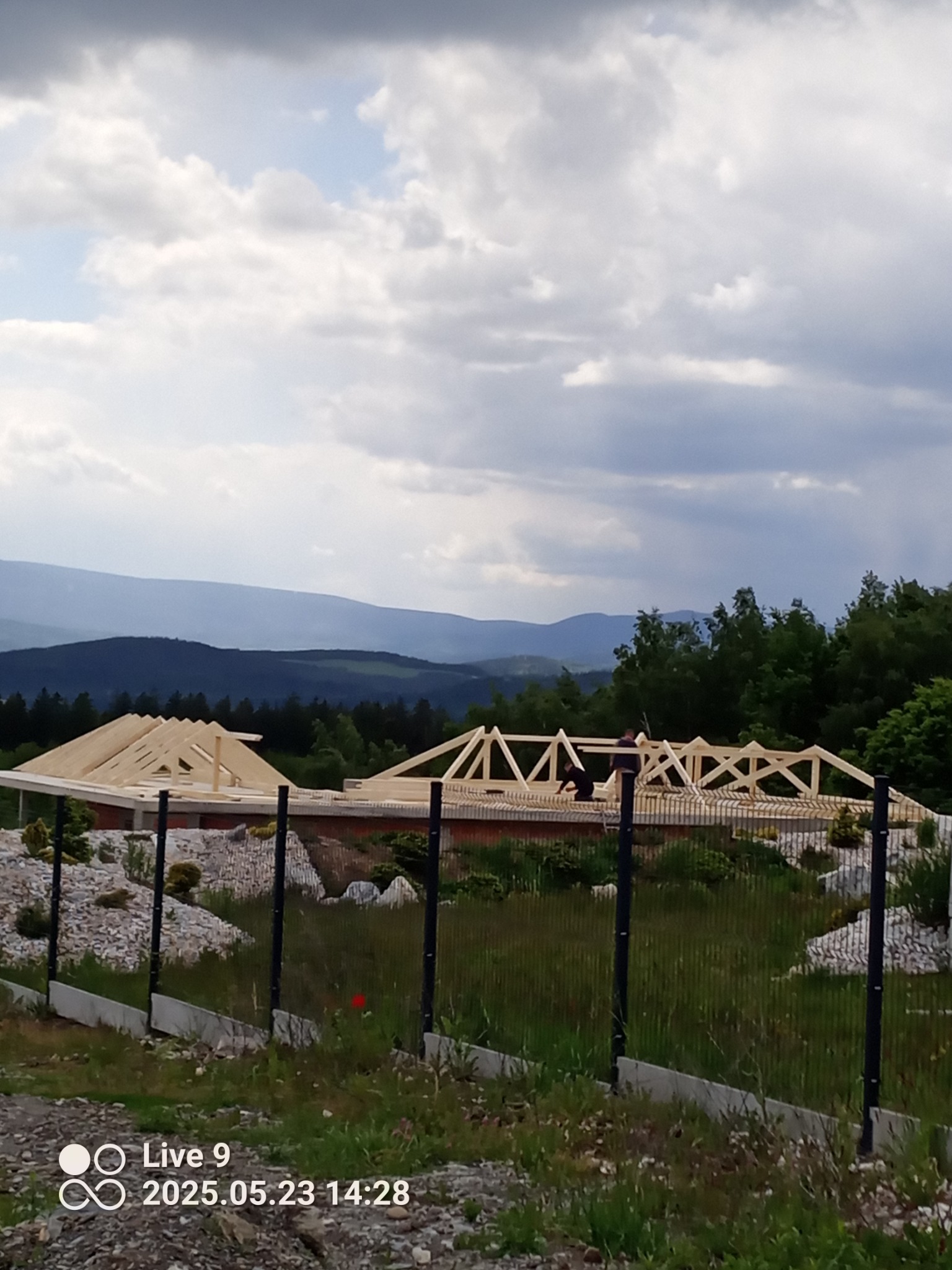 Wznoszenie więźby dachowej na murowanym domu jednorodzinnym w malowniczym krajobrazie. Widoczne elementy konstrukcyjne i pracownicy podczas montażu.