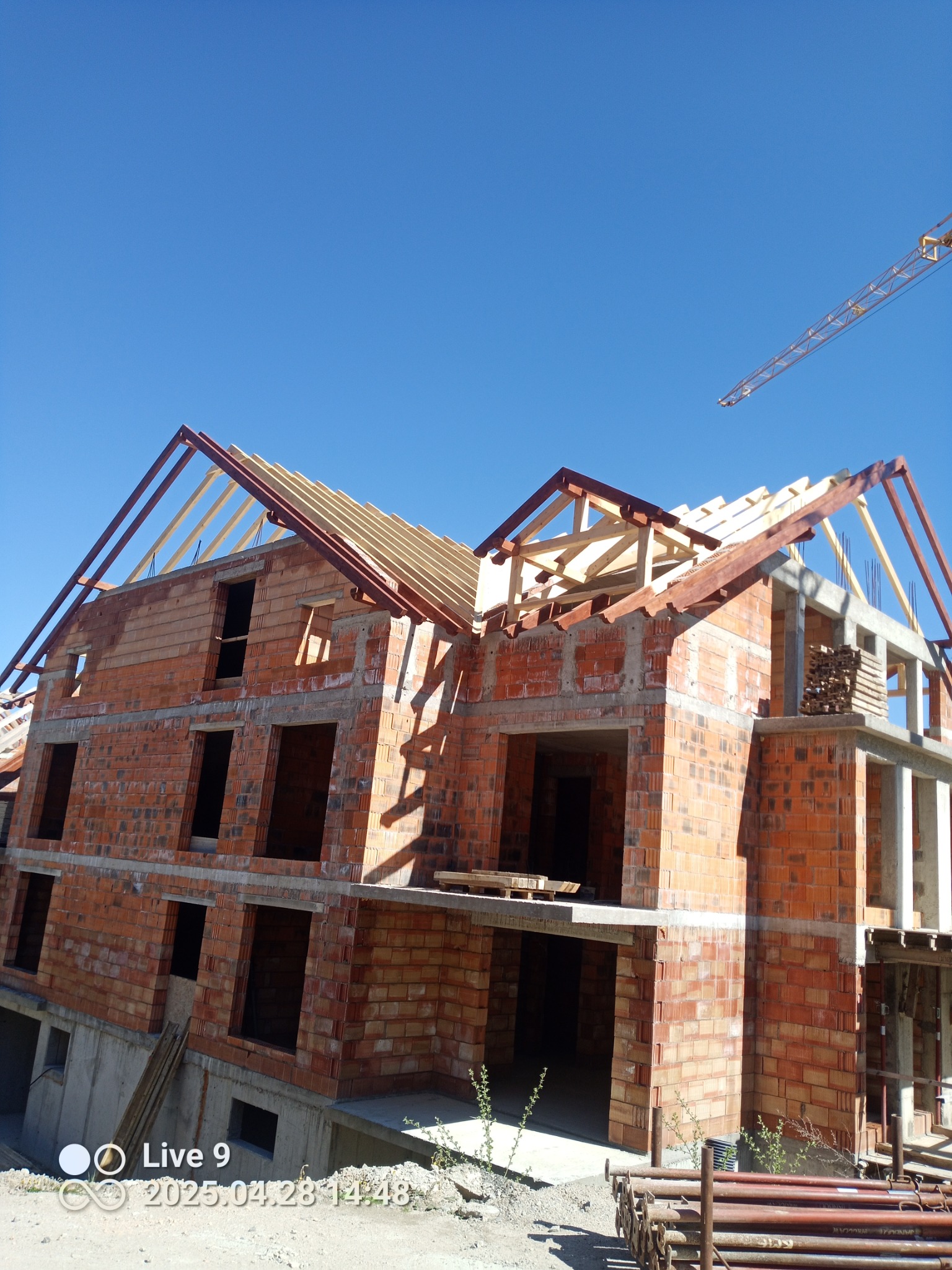 Wznoszenie surowego, dwupiętrowego budynku z czerwonej cegły, z częściowo ukończoną konstrukcją dachu z drewna, na tle błękitnego nieba i żurawia budowlanego.