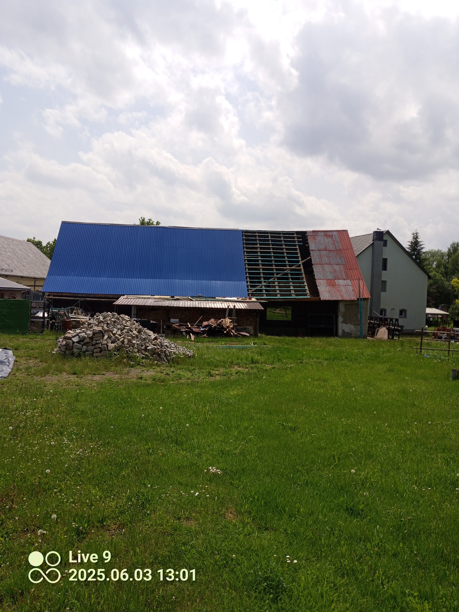 Remont dachu budynku gospodarczego: połowa pokryta blachą w kolorze niebieskim, druga połowa z odsłoniętą więźbą. Na pierwszym planie sterta kamieni.