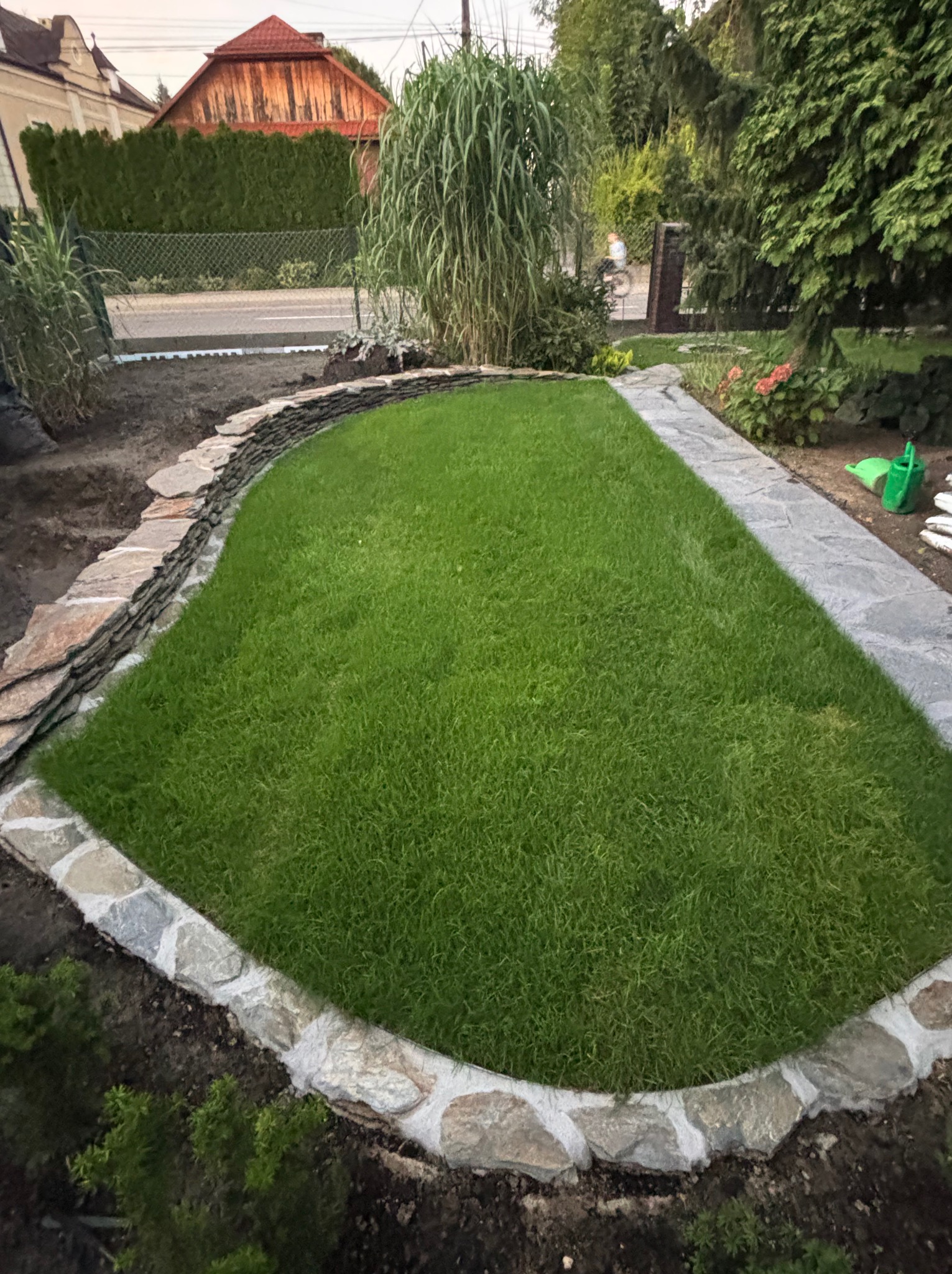 Zadbany trawnik otoczony kamiennym obrzeżem i ścieżką z płyt. W tle widoczna zieleń i zabudowania. Ujęcie z perspektywy lekko z góry, podkreślające kształt trawnika.