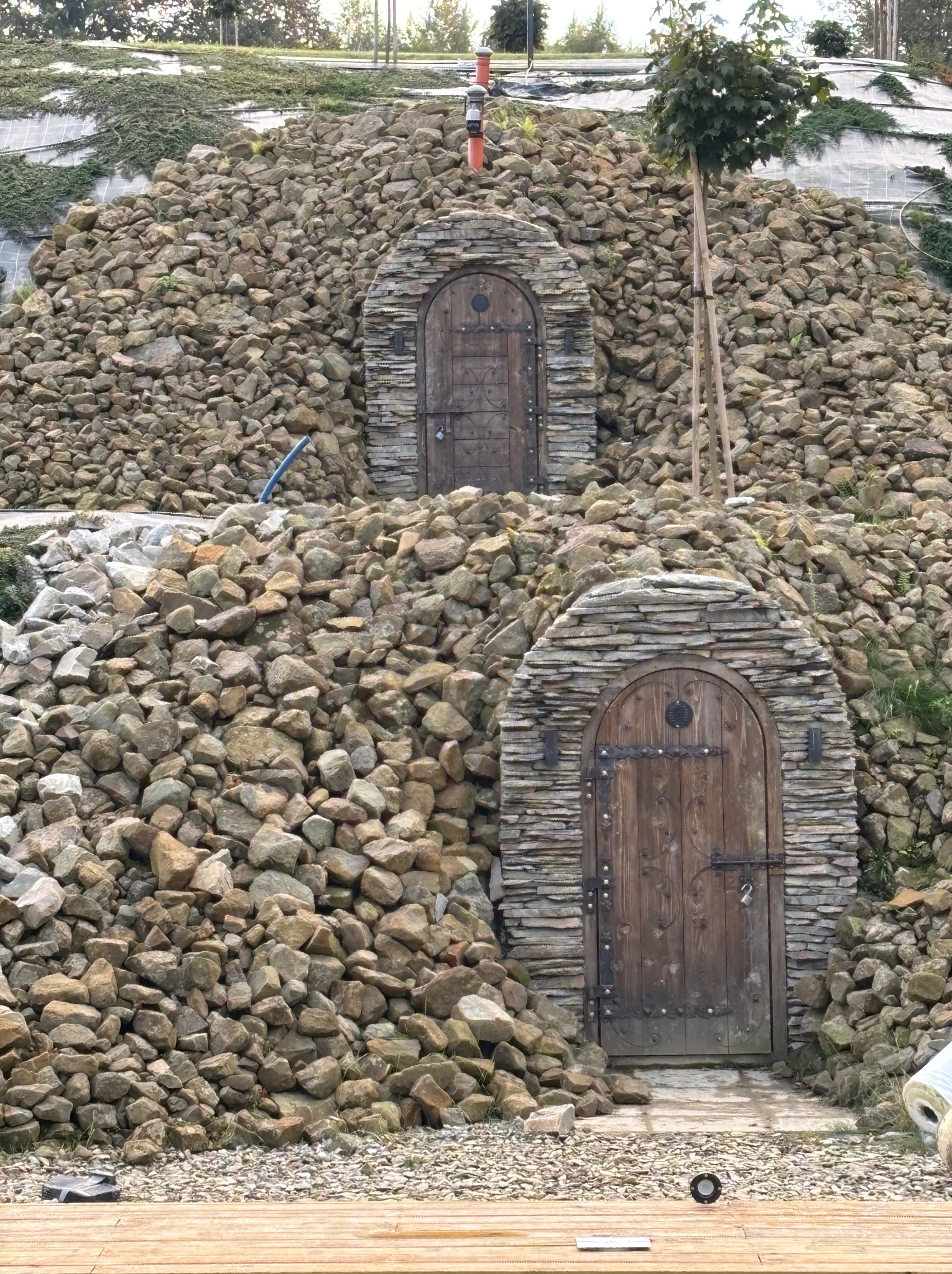 Dwa kamienne wejścia z drewnianymi drzwiami w zboczu wzgórza, otoczone kamieniami i roślinnością. Nietypowa architektura krajobrazu z elementami rustykalnymi.