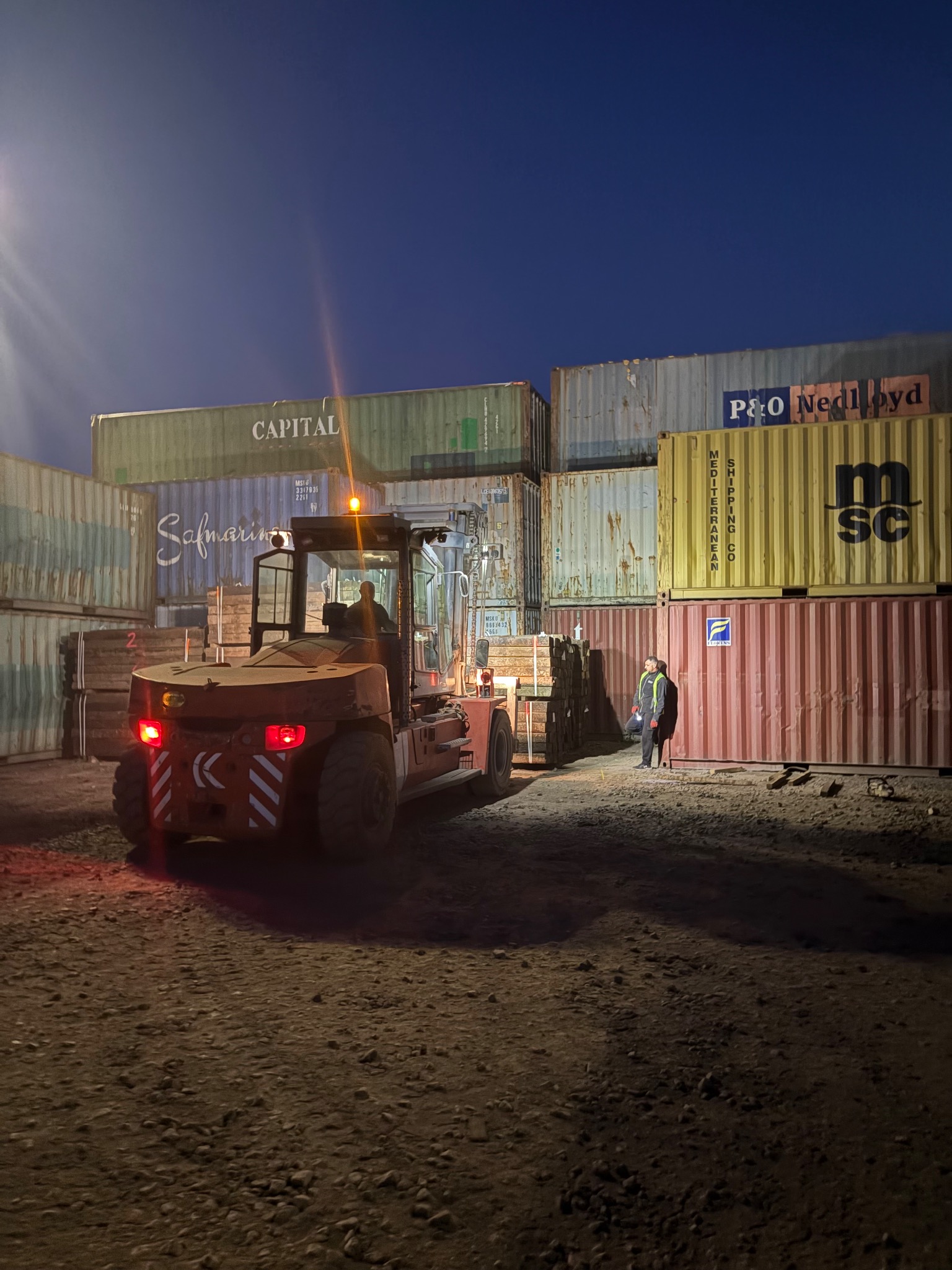 Wózek widłowy transportuje ładunek na tle kontenerów w porcie. Operator w kabinie, obok pracownik w kamizelce. Wieczorne oświetlenie.