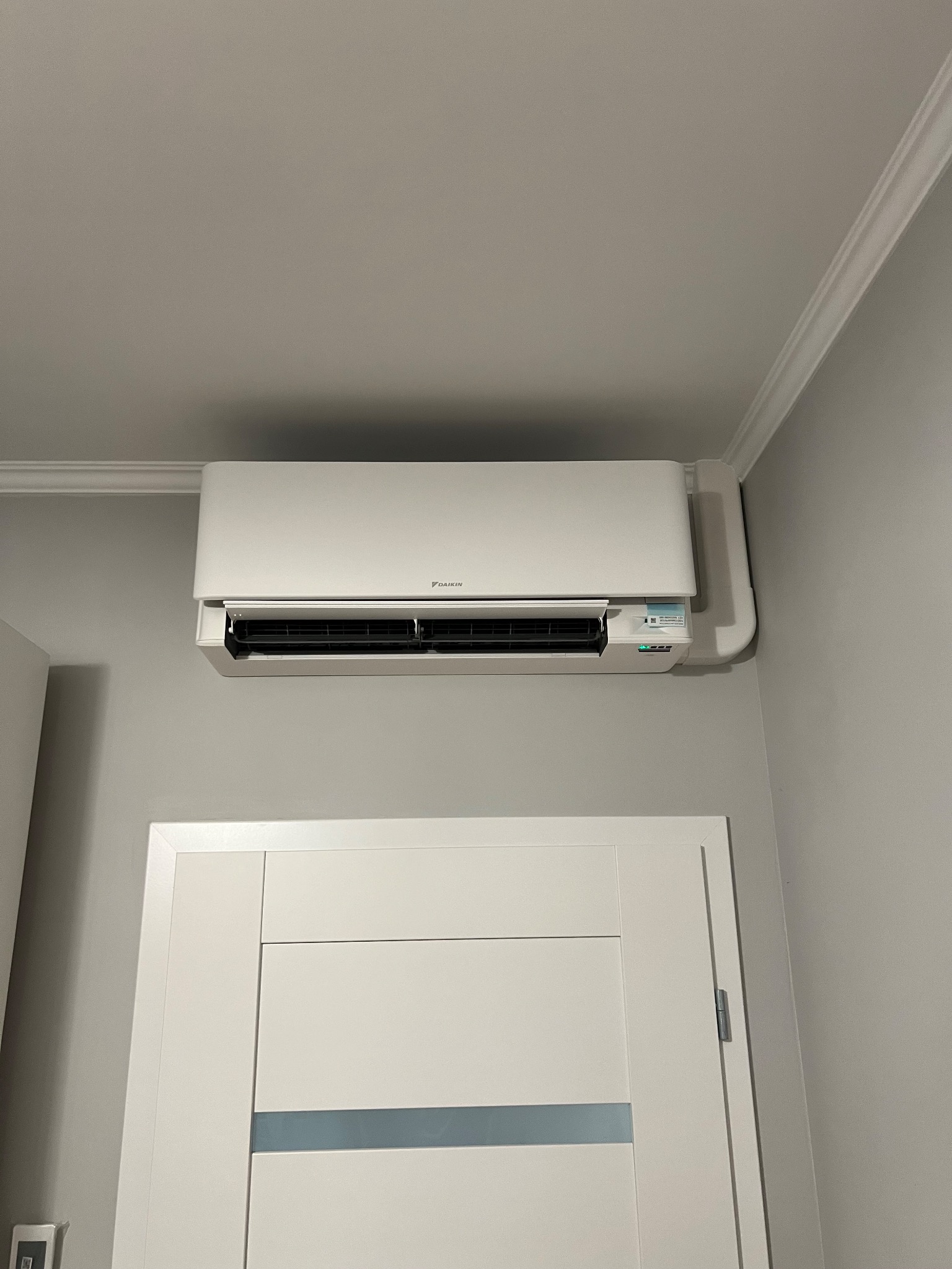 Biały klimatyzator Daikin zamontowany nad białą futryną drzwi w szarym pomieszczeniu. Widoczne listwy sufitowe.