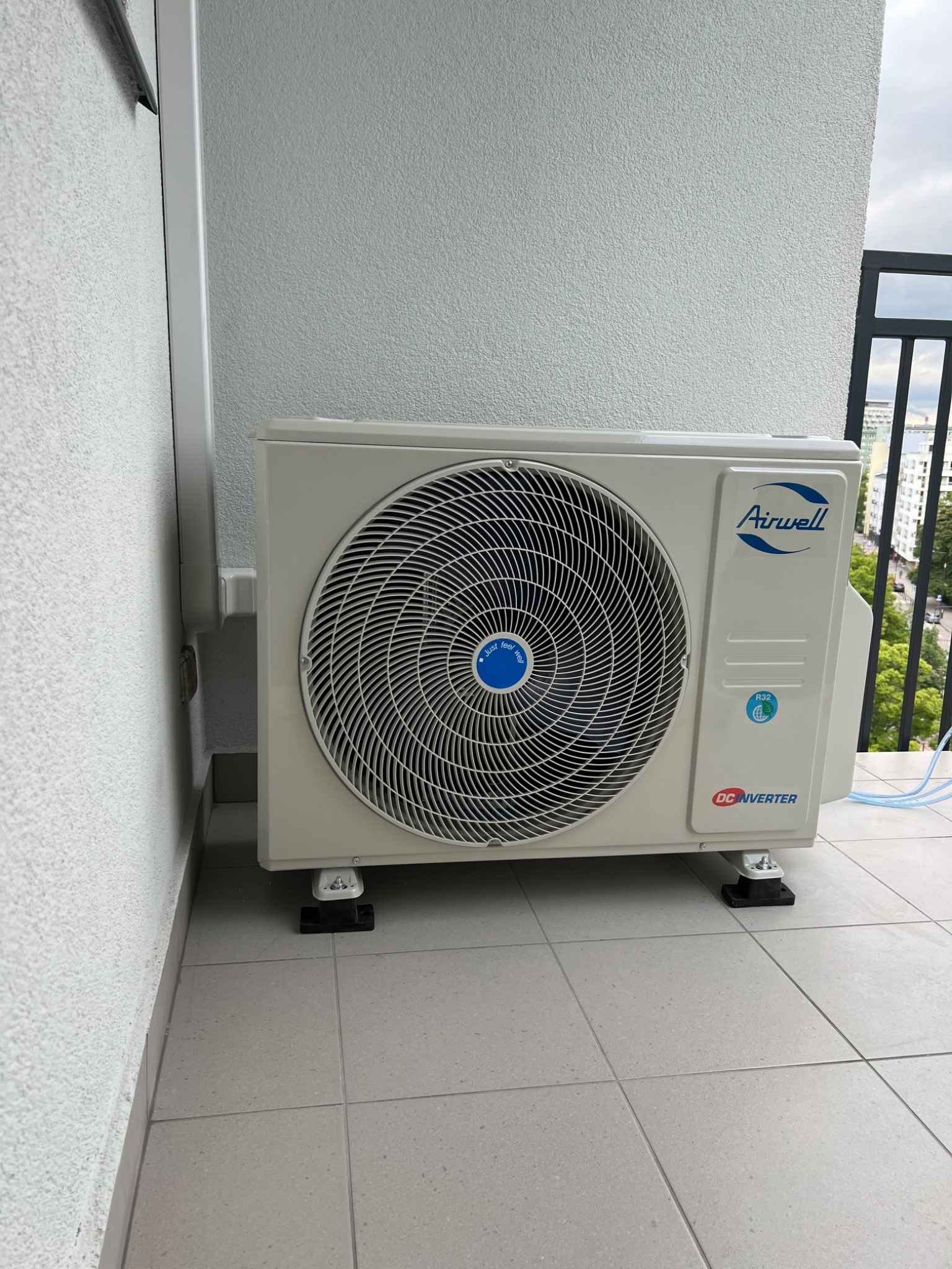 Zewnętrzna jednostka klimatyzacji Airwell na balkonie, z widocznymi przewodami i uchwytami, na tle miejskiego krajobrazu w pochmurny dzień.