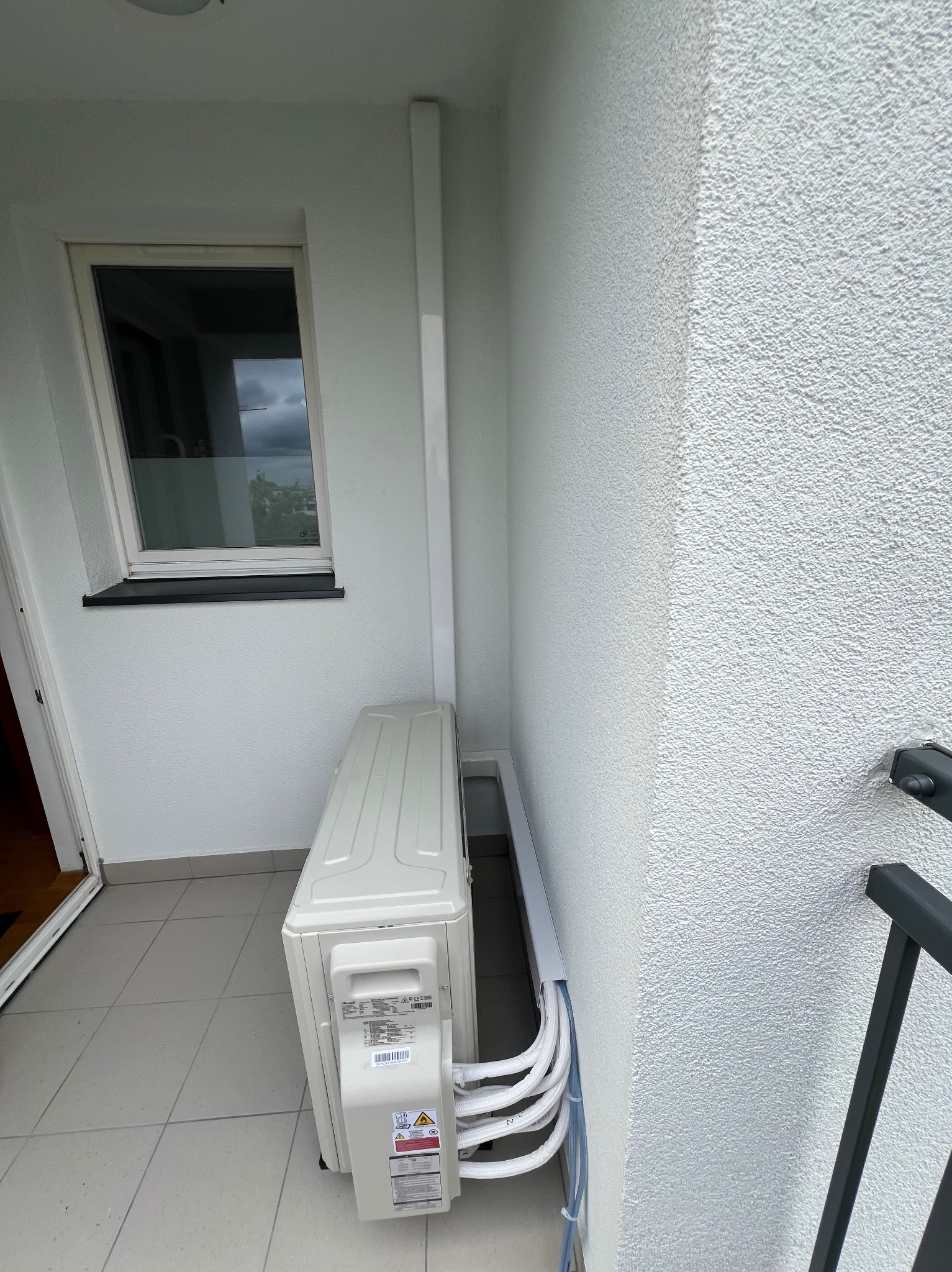 Biała jednostka zewnętrzna klimatyzacji na balkonie w Warszawie. Widoczne rury i kable prowadzące do wnętrza. Ściana z fakturą baranka.