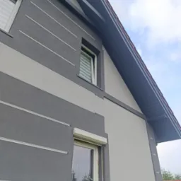 Dwukolorowa elewacja domu z szarymi pasami i fragment dachu z widoczną rynną dachową pod kątem, z roletami w oknach.