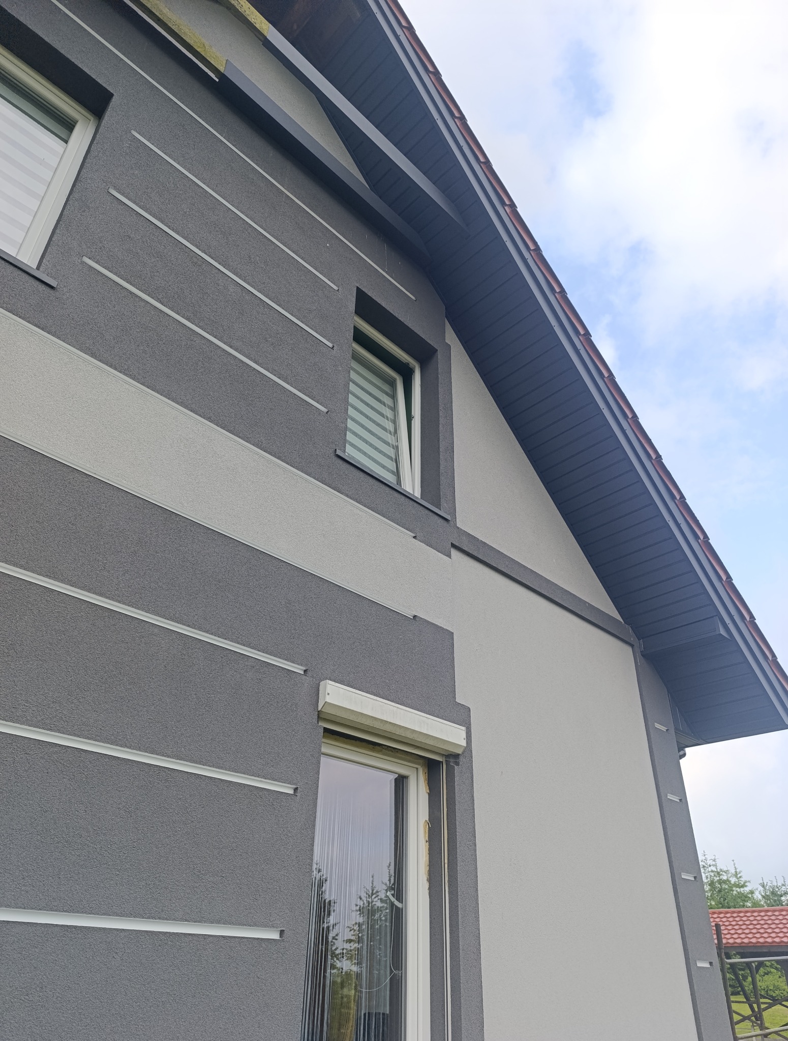 Dwukolorowa elewacja domu z szarymi pasami i fragment dachu z widoczną rynną dachową pod kątem, z roletami w oknach.