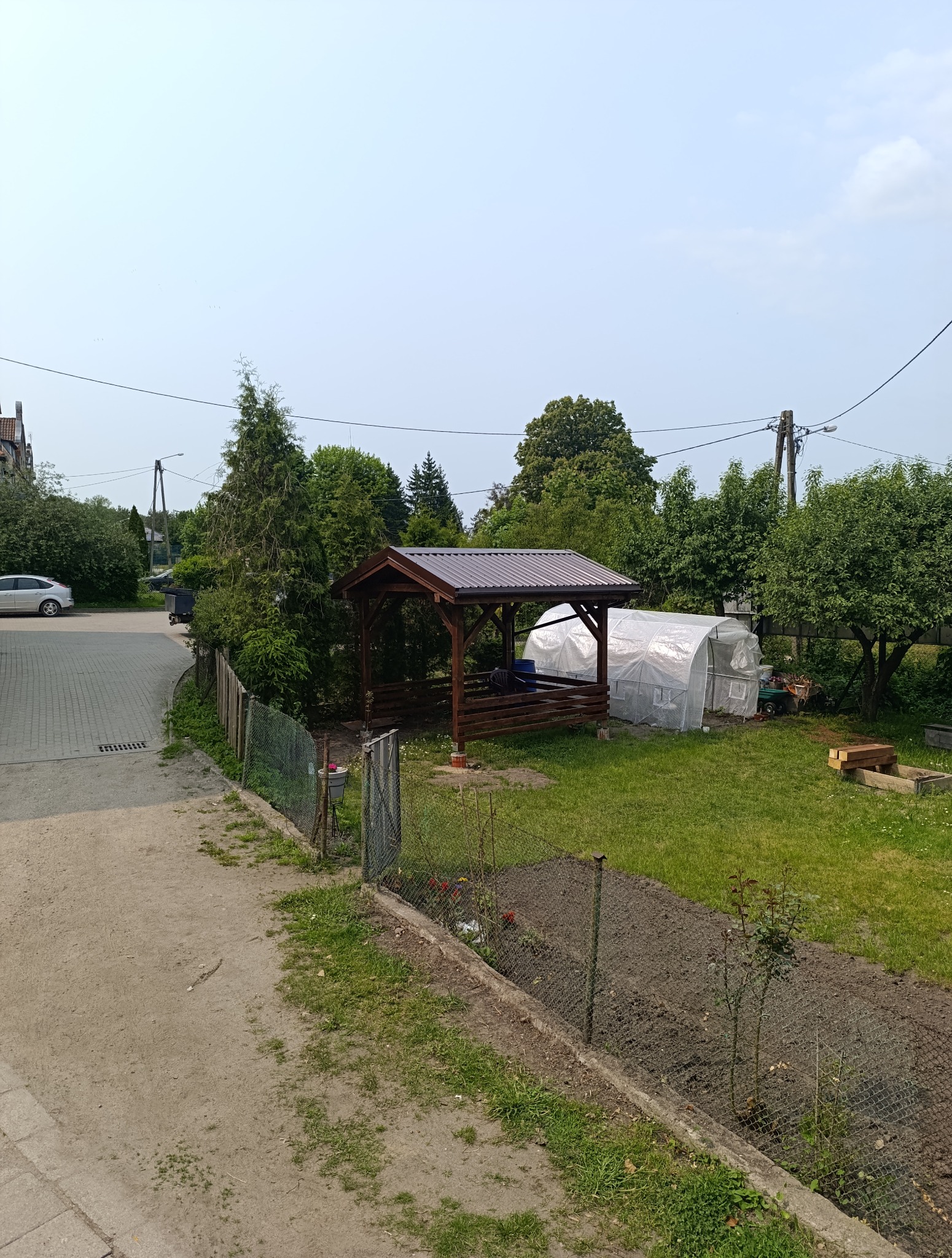 Pergola z brązowym dachem blaszany na zielonym trawniku, obok folia. Widok z góry na działkę z ogrodem i chodnikiem. Słoneczny dzień.