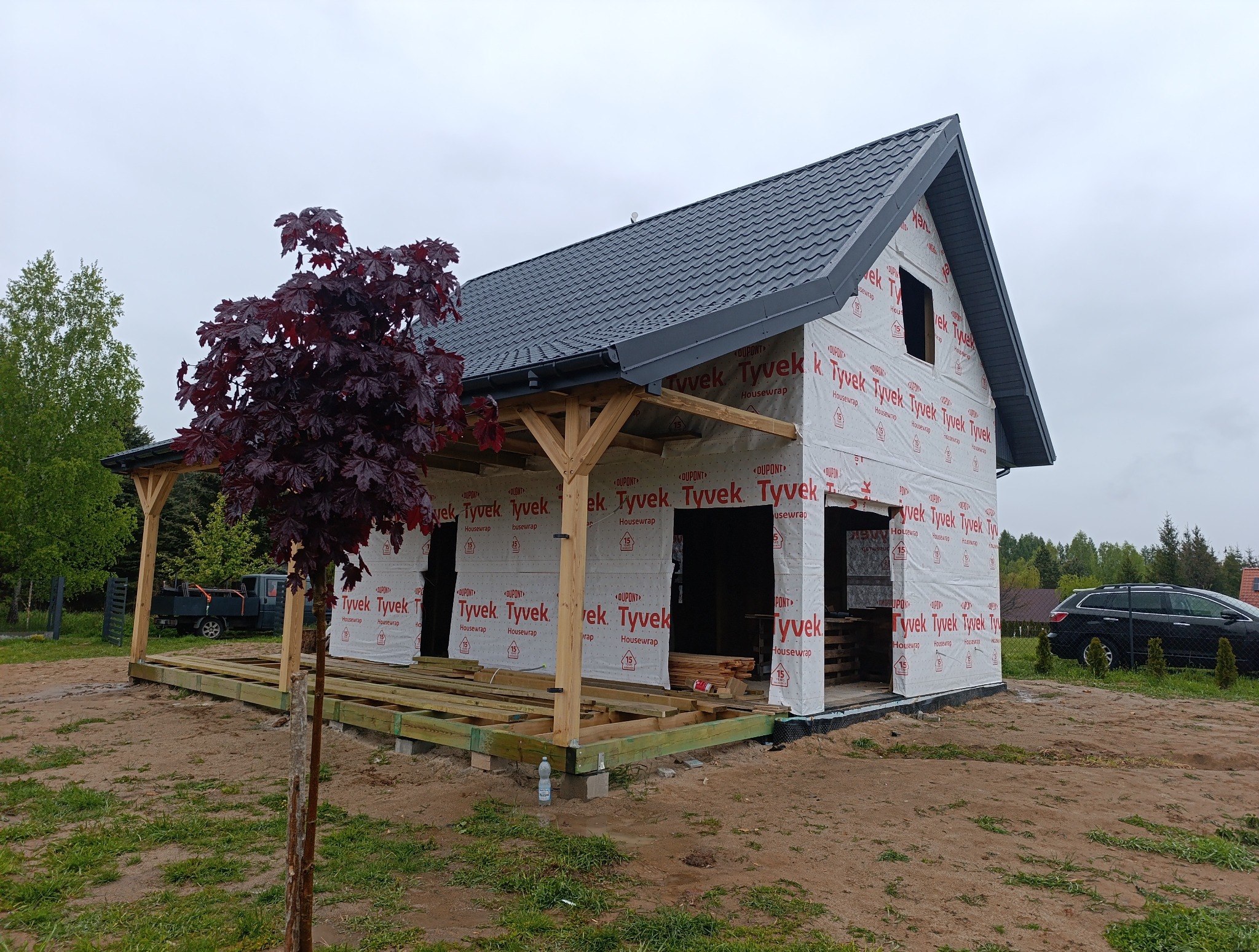 Dom w budowie z szarą dachówką, zabezpieczony membraną Tyvek, z drewnianą konstrukcją werandy i młodym drzewem klonu czerwonego na pierwszym planie.