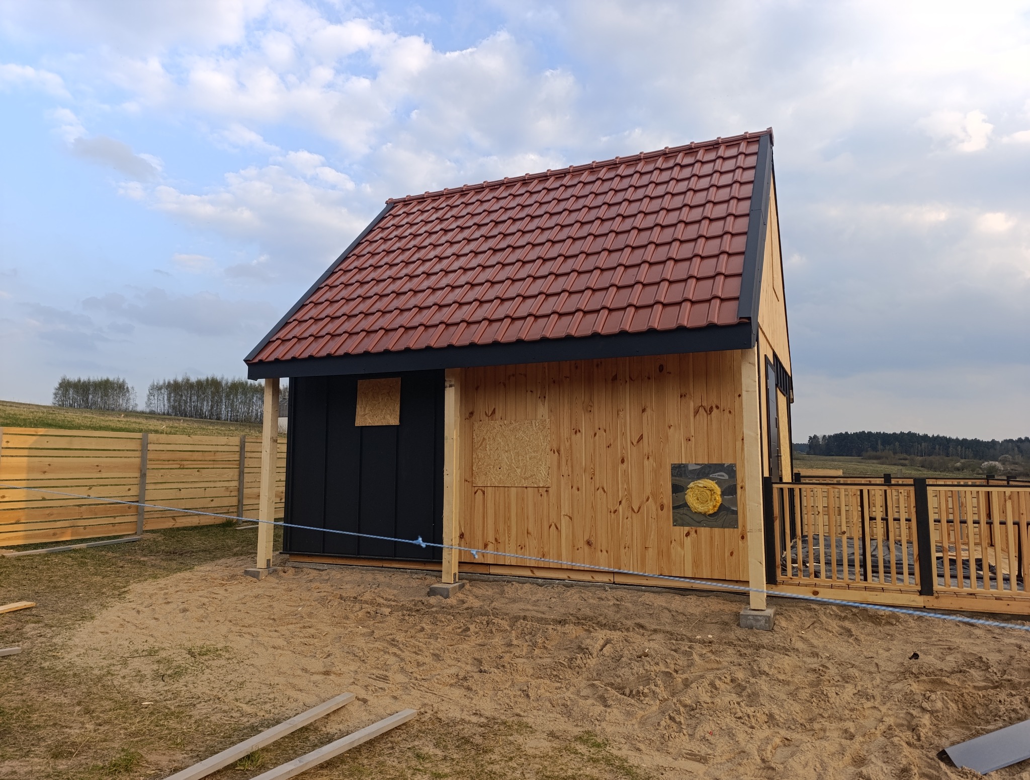 Niski budynek z brązową dachówką i drewnianą elewacją. Część ścian jest czarna. Okna zabite płytami. Wokół piasek i drewniane ogrodzenie. Widok na zalesione wzgórze.