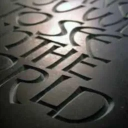 Wygrawerowany napis w ciemnym kamieniu, widoczne głębokie żłobienia liter w surowym materiale. Perspektywa z góry, oświetlenie boczne podkreśla teksturę.