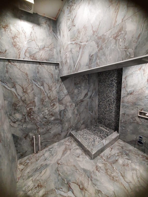 Wykończona kabina prysznicowa z szarymi płytkami imitującymi marmur na ścianach i podłodze. Mała mozaika wewnątrz kabiny. Widoczne rury.