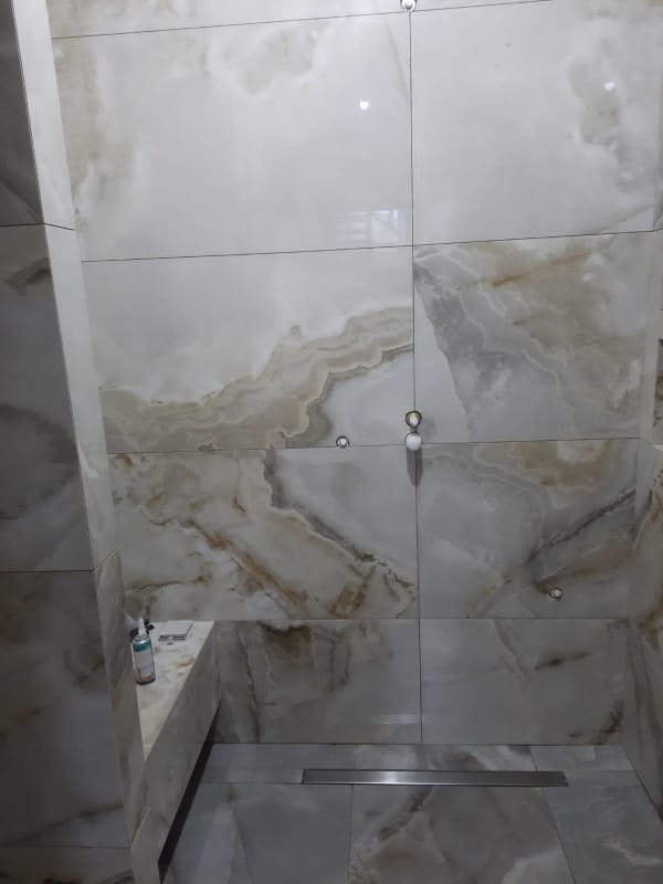 Wykończona łazienka z beżowymi płytkami imitującymi marmur. Widoczny odpływ liniowy i fragment umywalki z blatem. Realizacja po remoncie, z dbałością o detale.