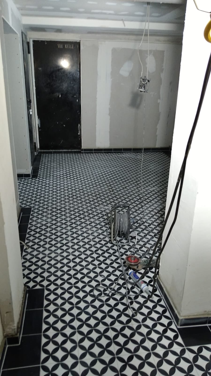 Wnętrze w trakcie wykończenia: świeżo położona, wzorzysta posadzka w czarno-białe romby, surowe ściany przygotowane do malowania, narzędzia pracy widoczne na podłodze.