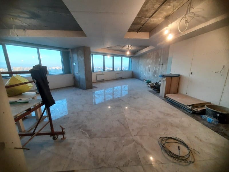 Wnętrze w trakcie wykańczania: marmurowe płytki podłogowe odbijają światło z okien z widokiem na miasto. Surowy beton na ścianach i suficie, widoczne instalacje elektryczne.