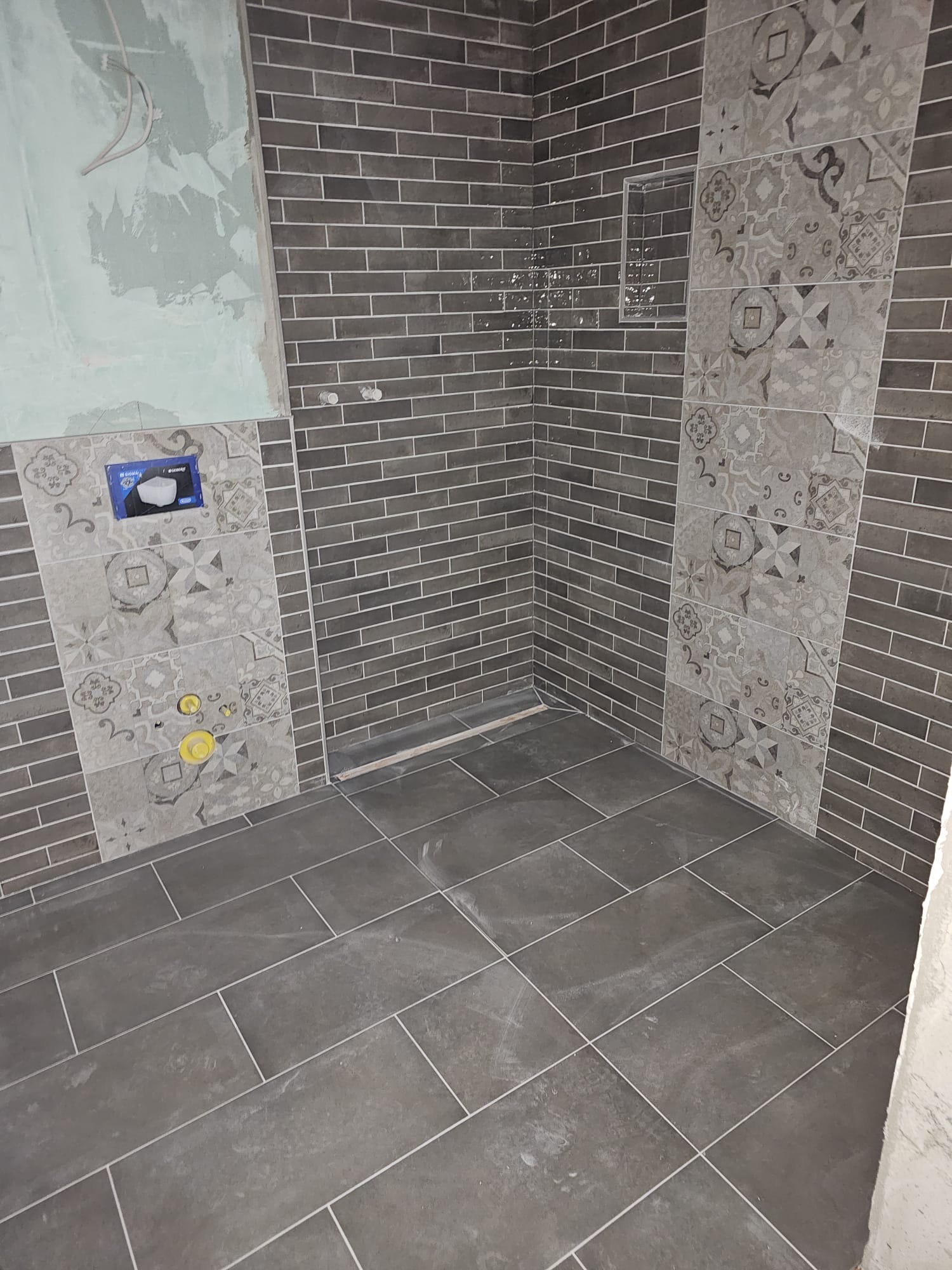 Szare płytki imitujące cegłę i dekoracyjne wnęki w łazience. Widoczne przygotowane instalacje pod armaturę.