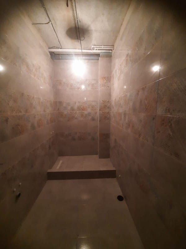 Wykończona łazienka z beżowymi płytkami w odcieniach brązu i kremu, widoczne rury pod sufitem. Minimalistyczny projekt, brak armatury.