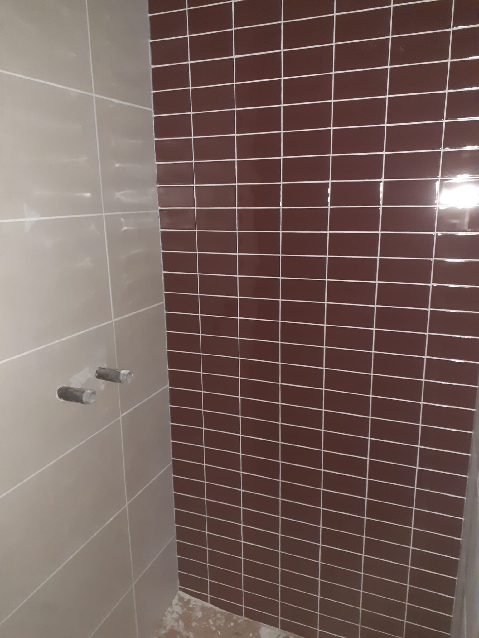 Wykończona ściana z brązowych, błyszczących płytek w małym formacie, z jasną fugą, sąsiadująca z jasną ścianą z widocznymi przyłączami hydraulicznymi.