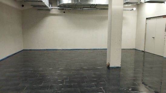 Wykończone wnętrze: ciemna, błyszcząca podłoga z płytek, białe ściany, kolumna. Widoczna taśma malarska przy podłodze. Sufit z instalacją wentylacyjną. Minimalistyczny styl.