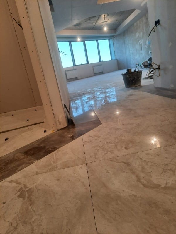 Wnętrze w trakcie wykończenia: marmurowe płytki podłogowe, ściany w stanie surowym, okna z widokiem. Widoczne narzędzia i materiały budowlane.