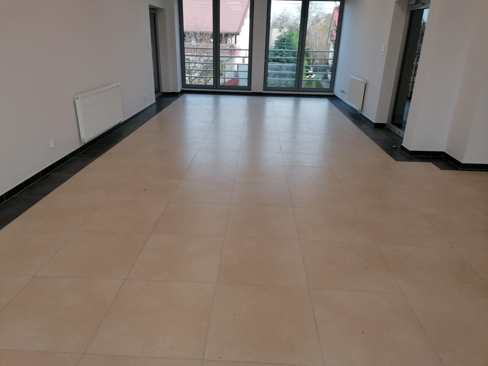 Jasna, przestronna sala z beżową podłogą z płytek, białymi ścianami i dużymi oknami balkonowymi z widokiem na zewnątrz. Grzejniki zamontowane na ścianach.