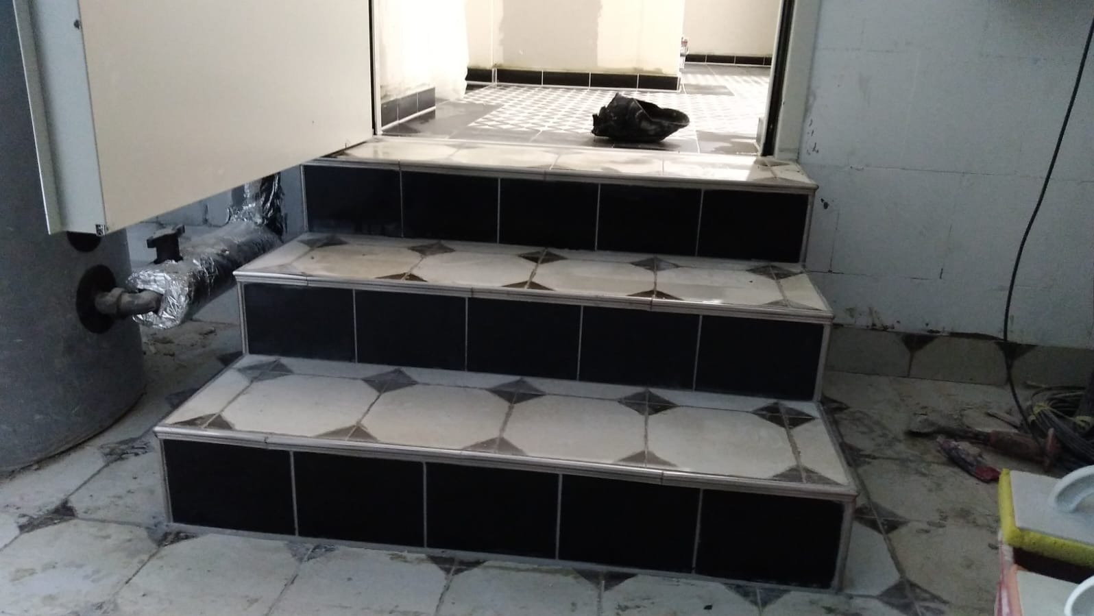 Wykończone płytkami schody z czarno-białym wzorem, prowadzące do pomieszczenia z podobną podłogą. Widoczny fragment pieca i instalacja. Prace wykończeniowe.