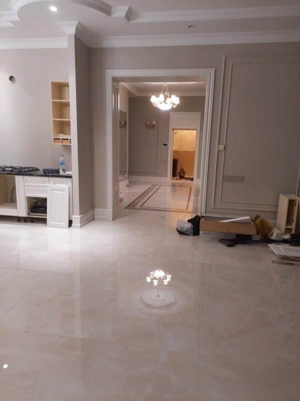 Eleganckie wnętrze w trakcie wykańczania: kremowe ściany, marmurowa podłoga z połyskiem, widoczne elementy zabudowy kuchennej i dekoracyjne sztukaterie.