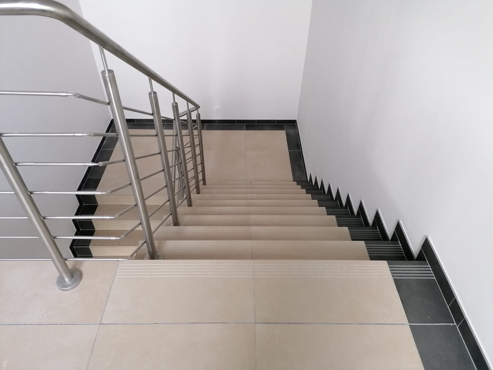 Wykończone schody z beżowymi stopniami i czarnymi płytkami przy ścianie. Metalowa, nowoczesna balustrada po lewej stronie. Widok z góry na dół.