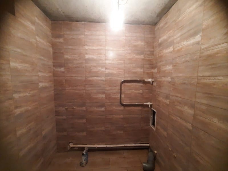 Wykończona łazienka z brązowymi płytkami imitującymi drewno na ścianach. Widoczne rury instalacji wodnej i kanalizacyjnej. Surowy betonowy sufit.