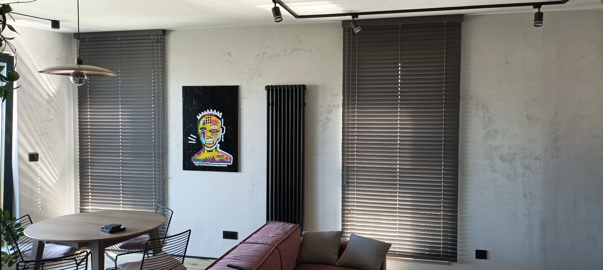 Szare żaluzje aluminiowe zamontowane w oknach nowoczesnego salonu z betonową ścianą, oświetleniem szynowym i obrazem w stylu Basquiata.