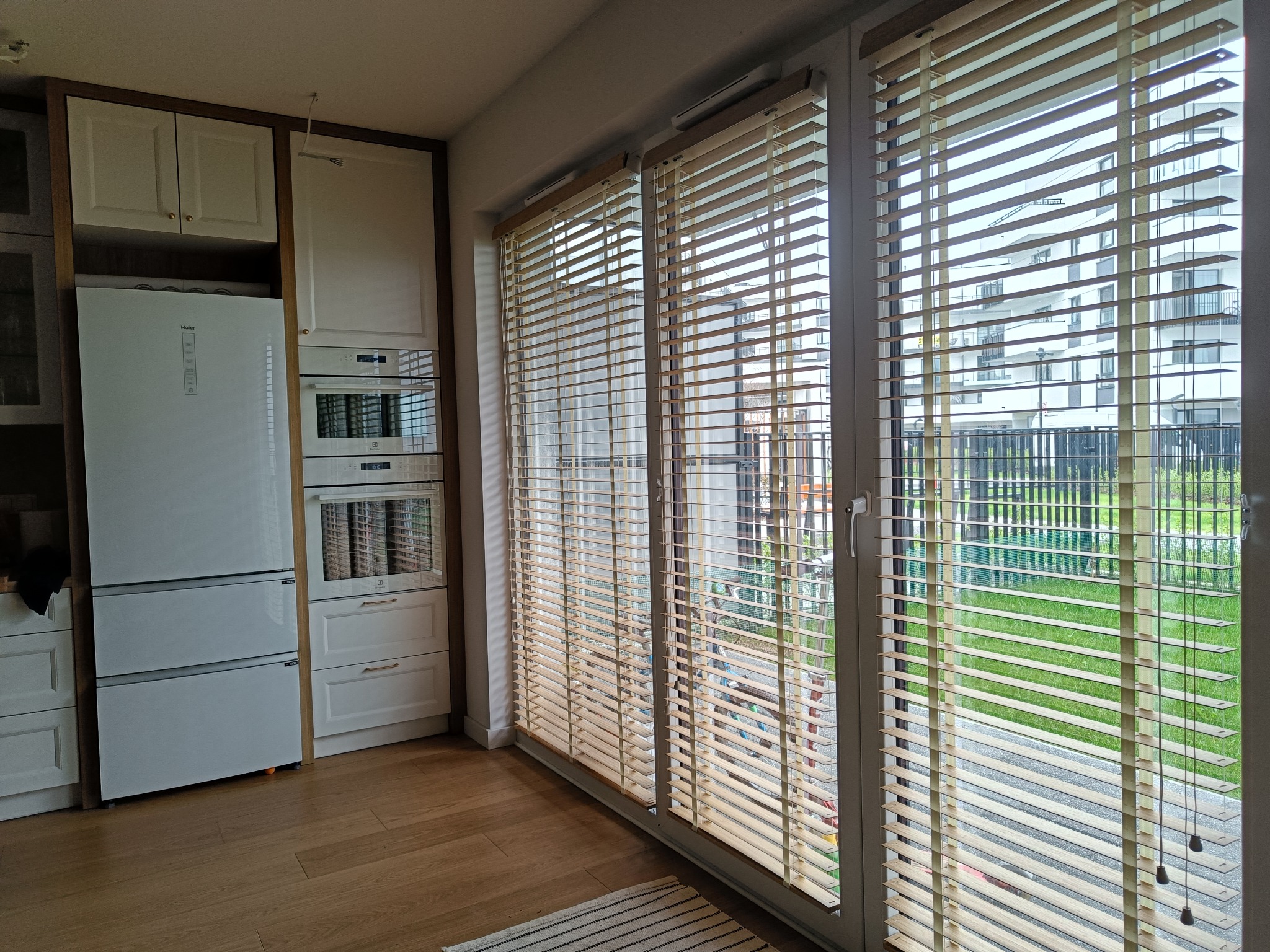 Jasne żaluzje drewniane na oknie balkonowym w kuchni. Widok na osiedle przez częściowo otwarte lamele. Minimalistyczny design, ciepłe odcienie drewna.