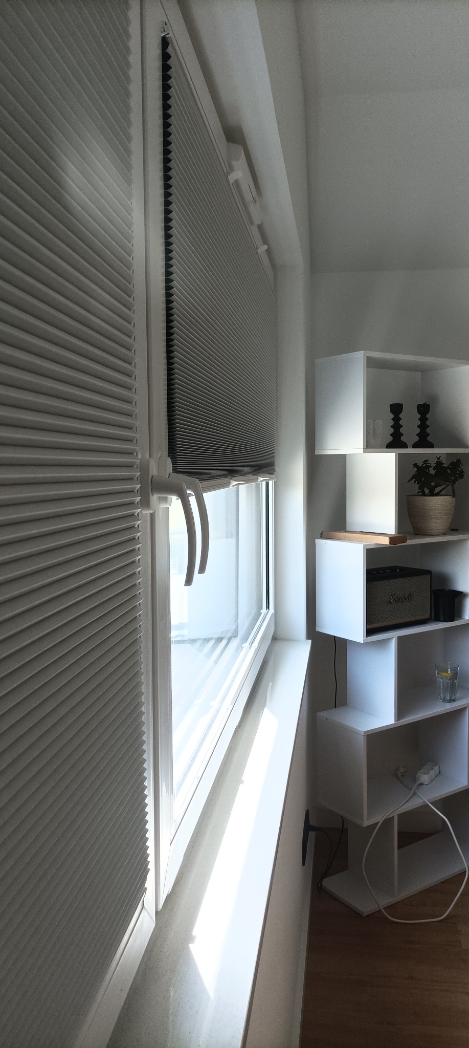 Jasna, plisowana osłona okienna zamontowana w oknie, z widocznymi uchwytami. Obok biały regał z dekoracjami.