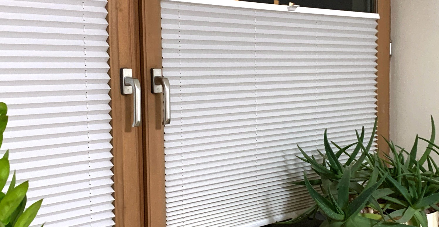 Zamknięte, białe plisy okienne w drewnianej ramie, z uchwytami. W tle i na pierwszym planie rośliny doniczkowe. Delikatne, rozproszone światło.