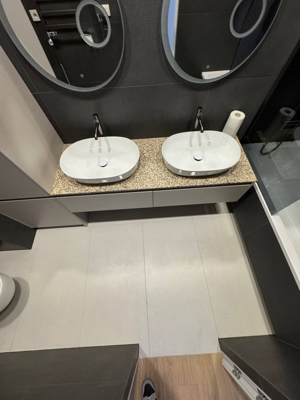 Nowoczesna łazienka z dwoma owalnymi umywalkami, lustrami z podświetleniem LED i szarą zabudową. Minimalistyczny design z płytkami podłogowymi w odcieniach szarości.