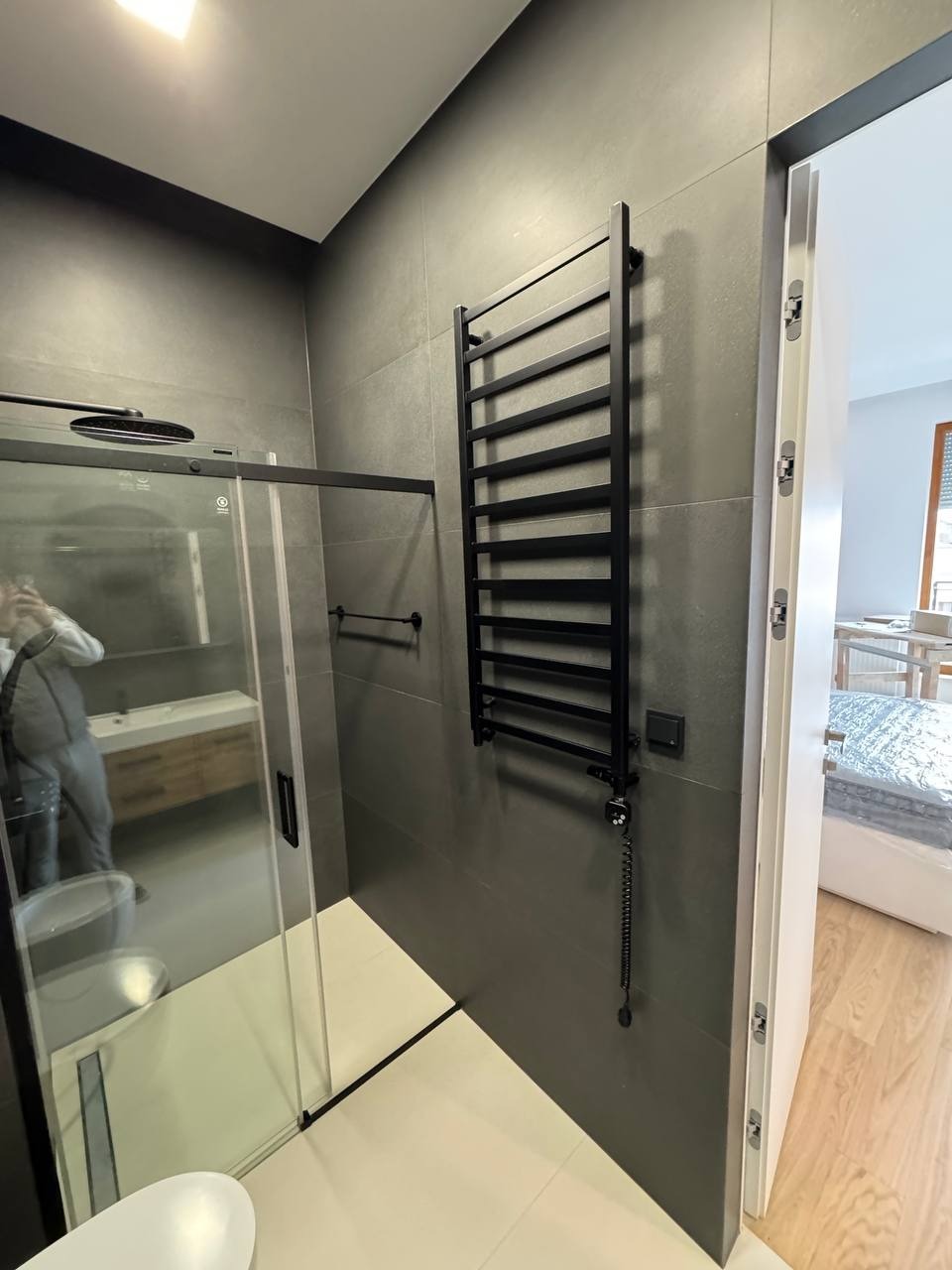 Nowoczesna łazienka z kabiną prysznicową, grzejnikiem drabinkowym i szarymi płytkami. Widoczny fragment toalety i odbicie osoby robiącej zdjęcie w szkle kabiny.