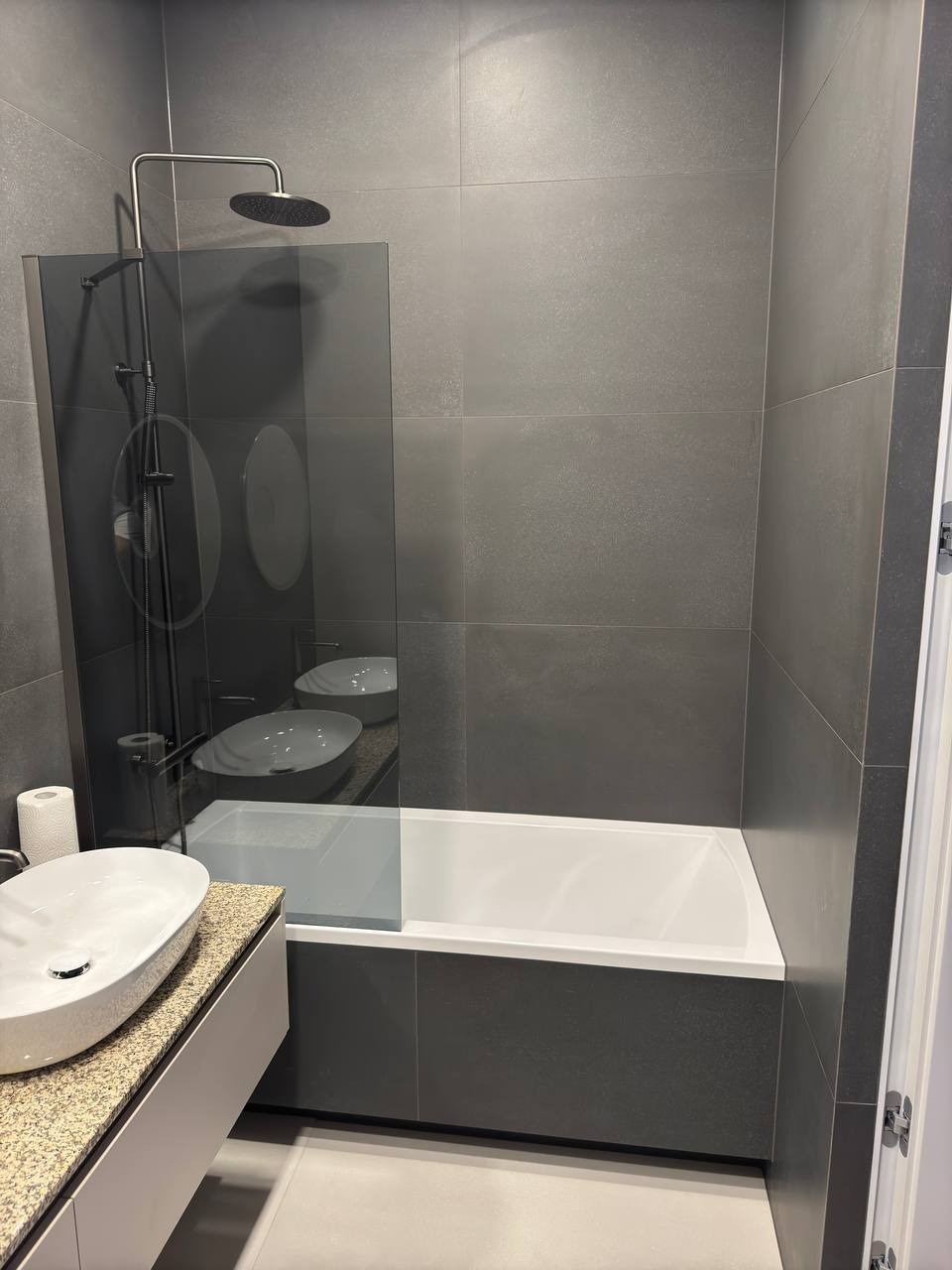 Nowoczesna łazienka w szarościach: biała wanna z parawanem, umywalka na blacie z kamienia. Minimalistyczny design, duże płytki, elegancka armatura.