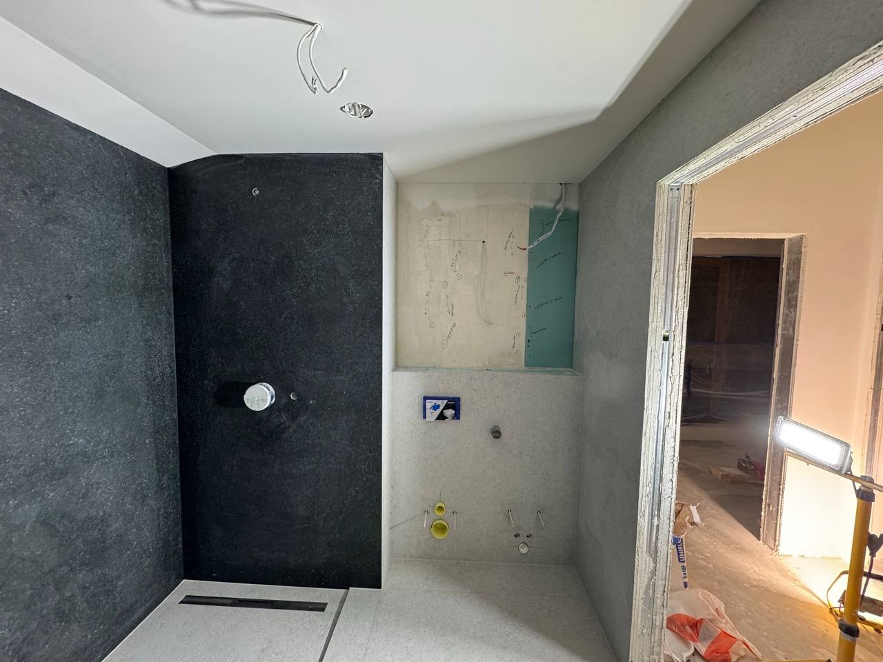 Wnętrze łazienki w trakcie wykończenia w Warszawie. Widoczna czarna ściana prysznicowa, białe płytki i instalacje. Prace wykończeniowe w toku, surowy stan.