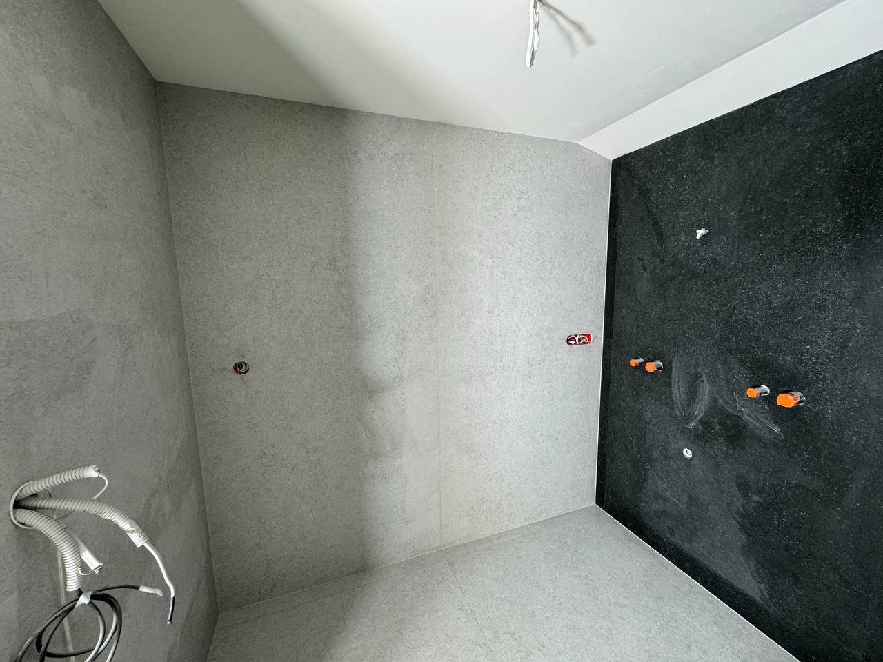 Wykończona łazienka z szarymi i czarnymi płytkami. Widoczne instalacje wodne i elektryczne. Minimalistyczny design, surowe wykończenie.