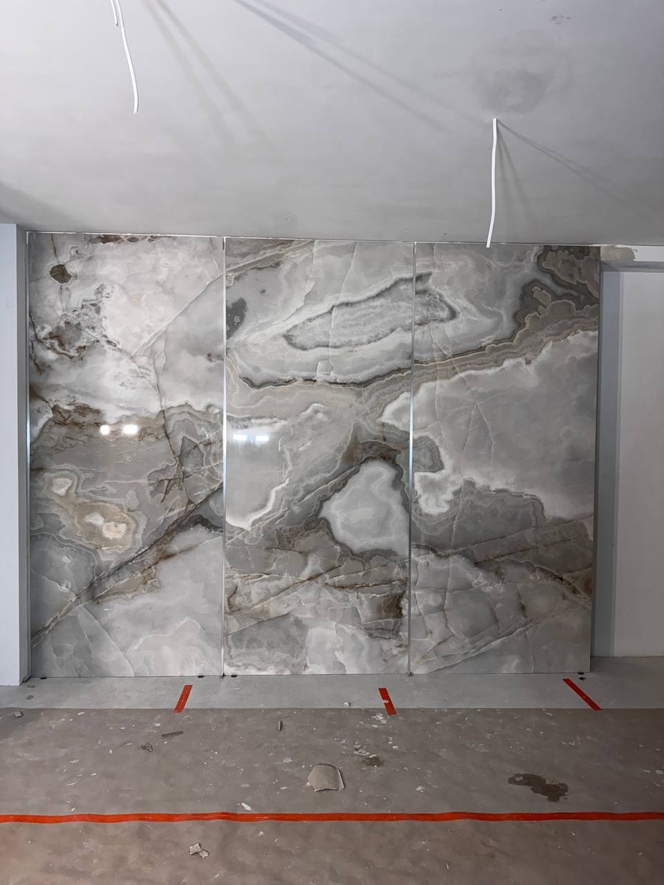 Ściana wyłożona trzema dużymi płytami z kamienia onyksowego o unikalnym, marmurkowym wzorze w odcieniach szarości i bieli, z widocznymi spoinami i fragmentem surowego pomieszczenia.