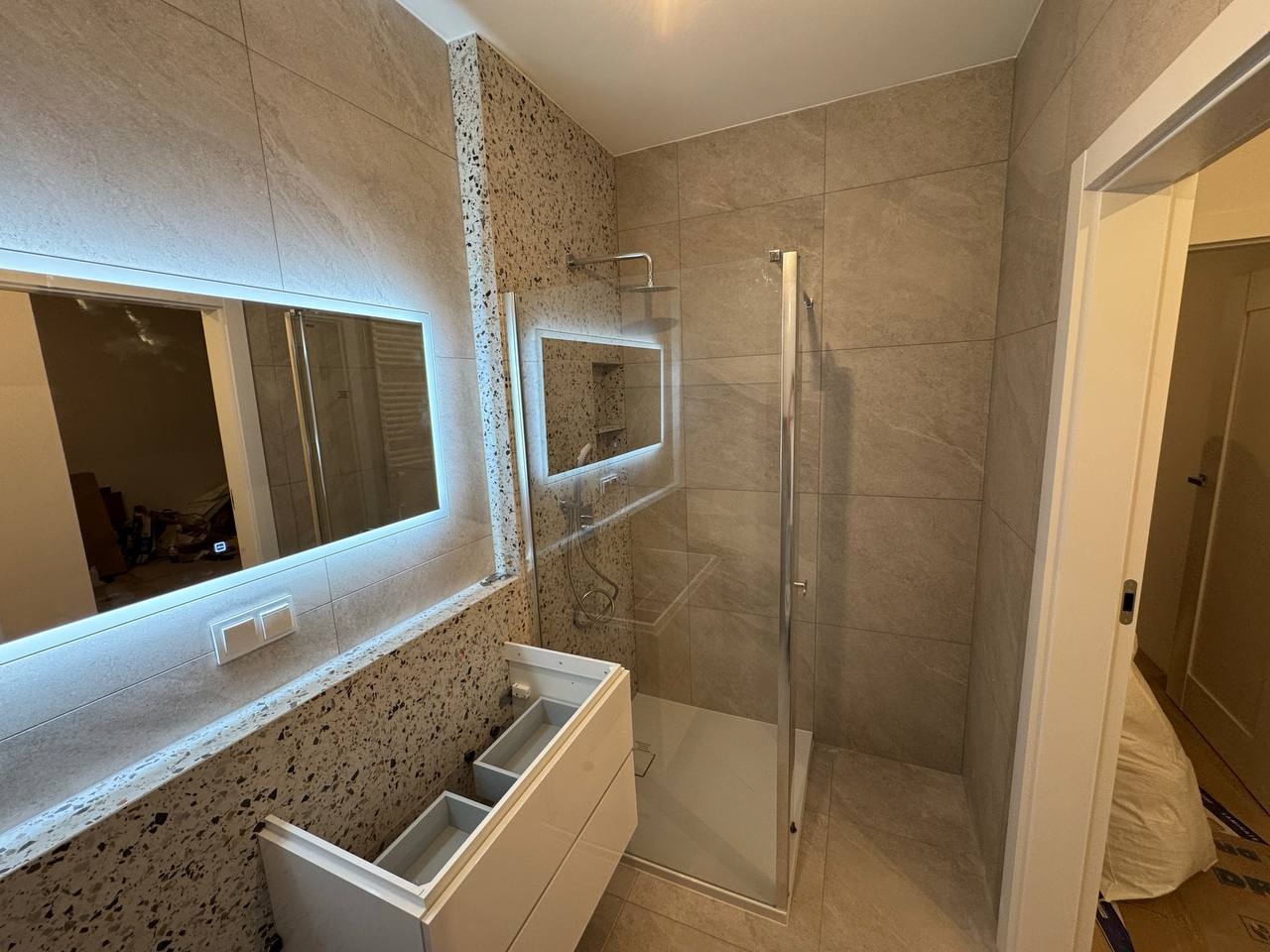 Nowoczesna łazienka w odcieniach szarości z podświetlanym lustrem i kabiną prysznicową. Umywalka bez frontów, widoczne wnętrze szafki. Wykończenie ścian imitujące lastryko.