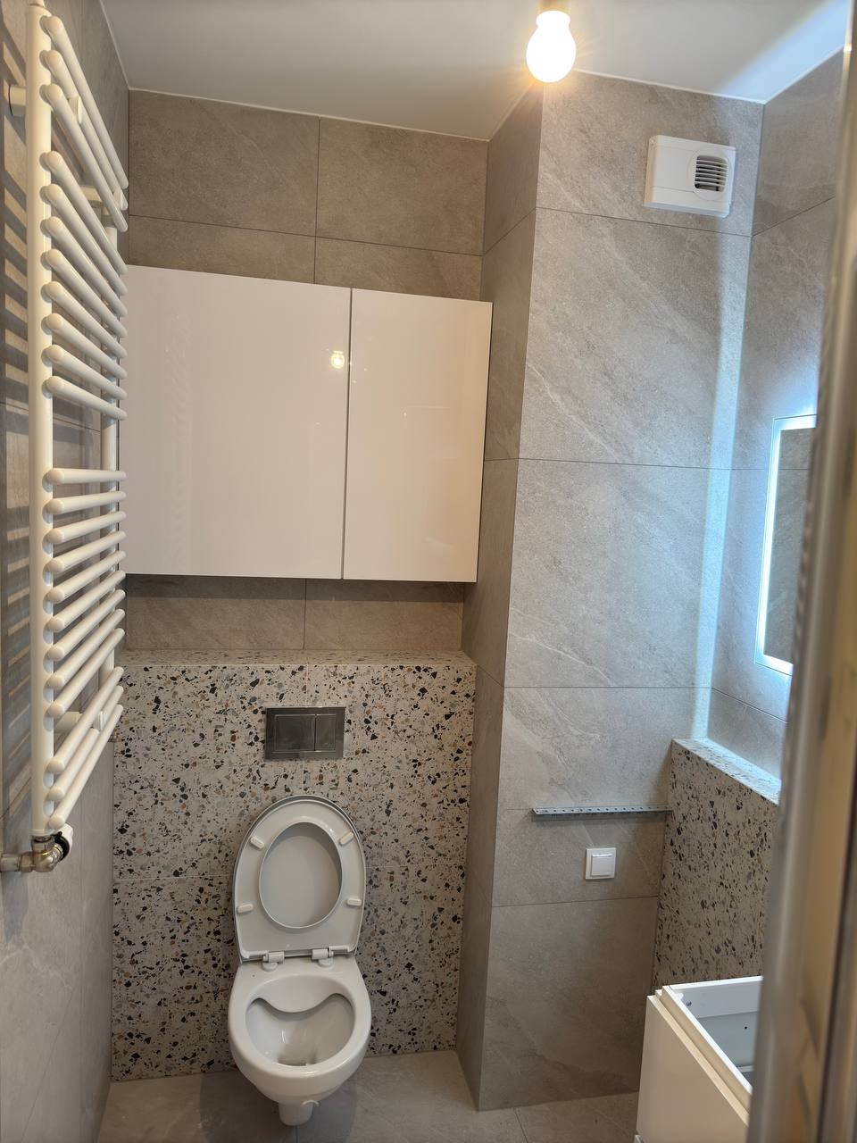 Wykończona łazienka z szarymi płytkami imitującymi kamień, toaleta z otwartą klapą, biała szafka wisząca i grzejnik drabinkowy. Nowoczesny design.