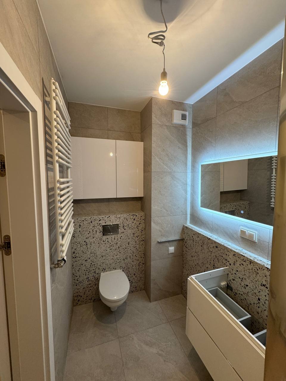 Nowoczesna łazienka z szarą płytką, podświetlanym lustrem i wiszącą szafką. Uwagę zwraca nietypowa ściana z mozaiką w strefie WC i umywalki.