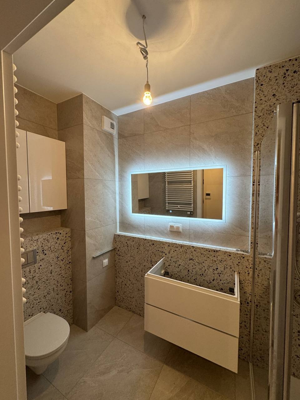 Jasna łazienka z podświetlanym lustrem, płytkami imitującymi kamień i niedokończoną szafką pod umywalkę. Widoczna toaleta i kabina prysznicowa.