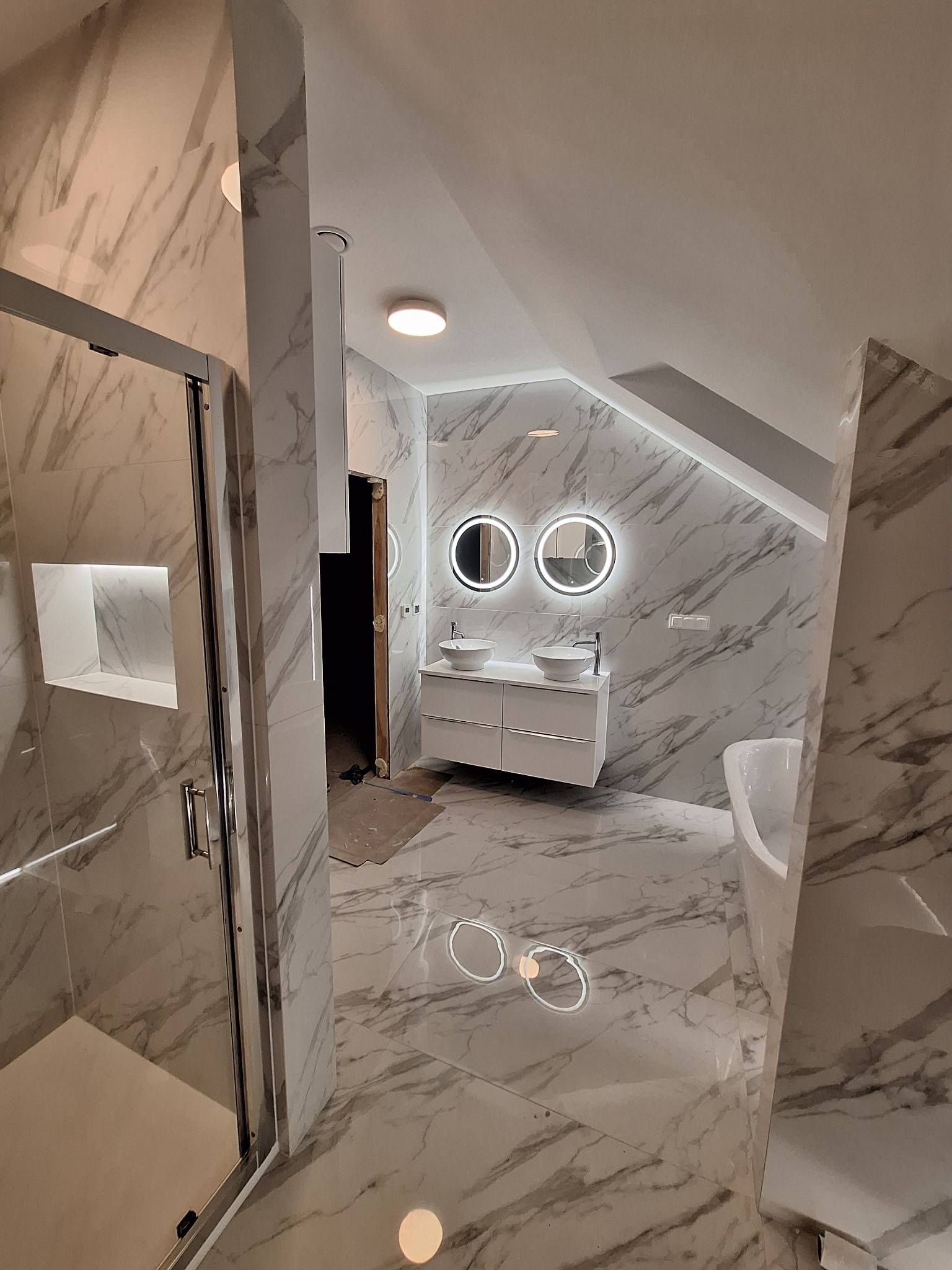 Elegancka łazienka na poddaszu z marmurowymi płytkami, podświetlanymi lustrami i kabiną prysznicową. Umywalki nablatowe i wanna wkomponowane w przestrzeń.