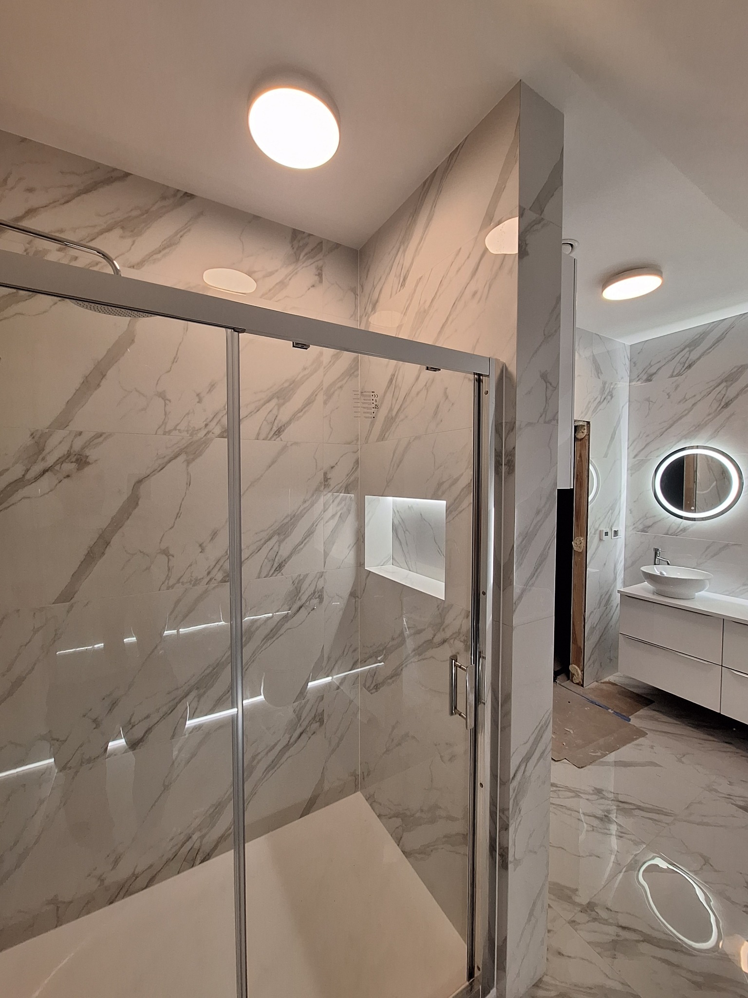 Wykończona łazienka z kabiną prysznicową typu walk-in, marmurowe płytki na ścianach i podłodze, oświetlenie LED i okrągłe lustro z podświetleniem.