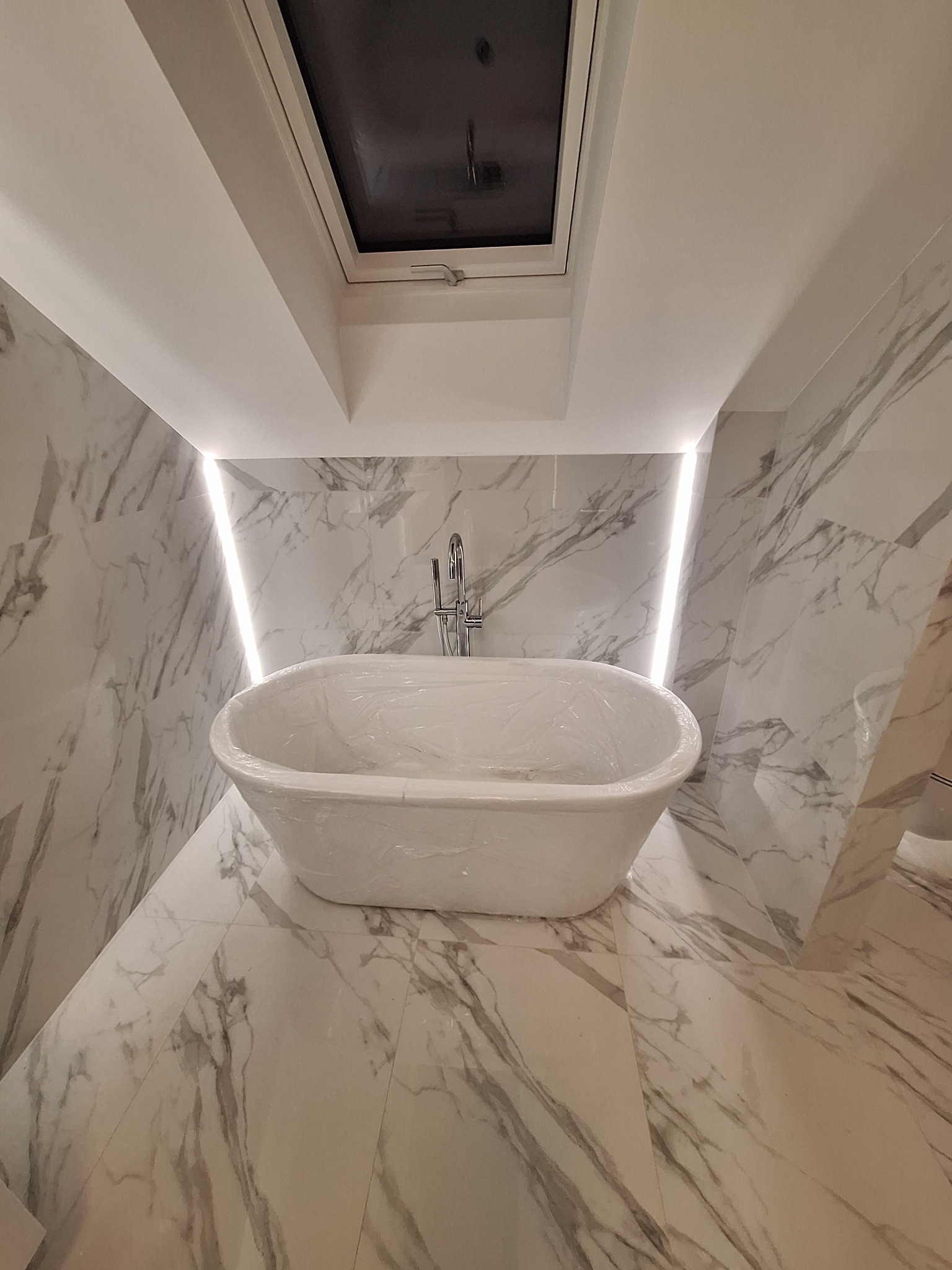 Nowoczesna łazienka z wolnostojącą wanną zabezpieczoną folią, marmurowe płytki na ścianach i podłodze, oświetlenie LED i okno dachowe.