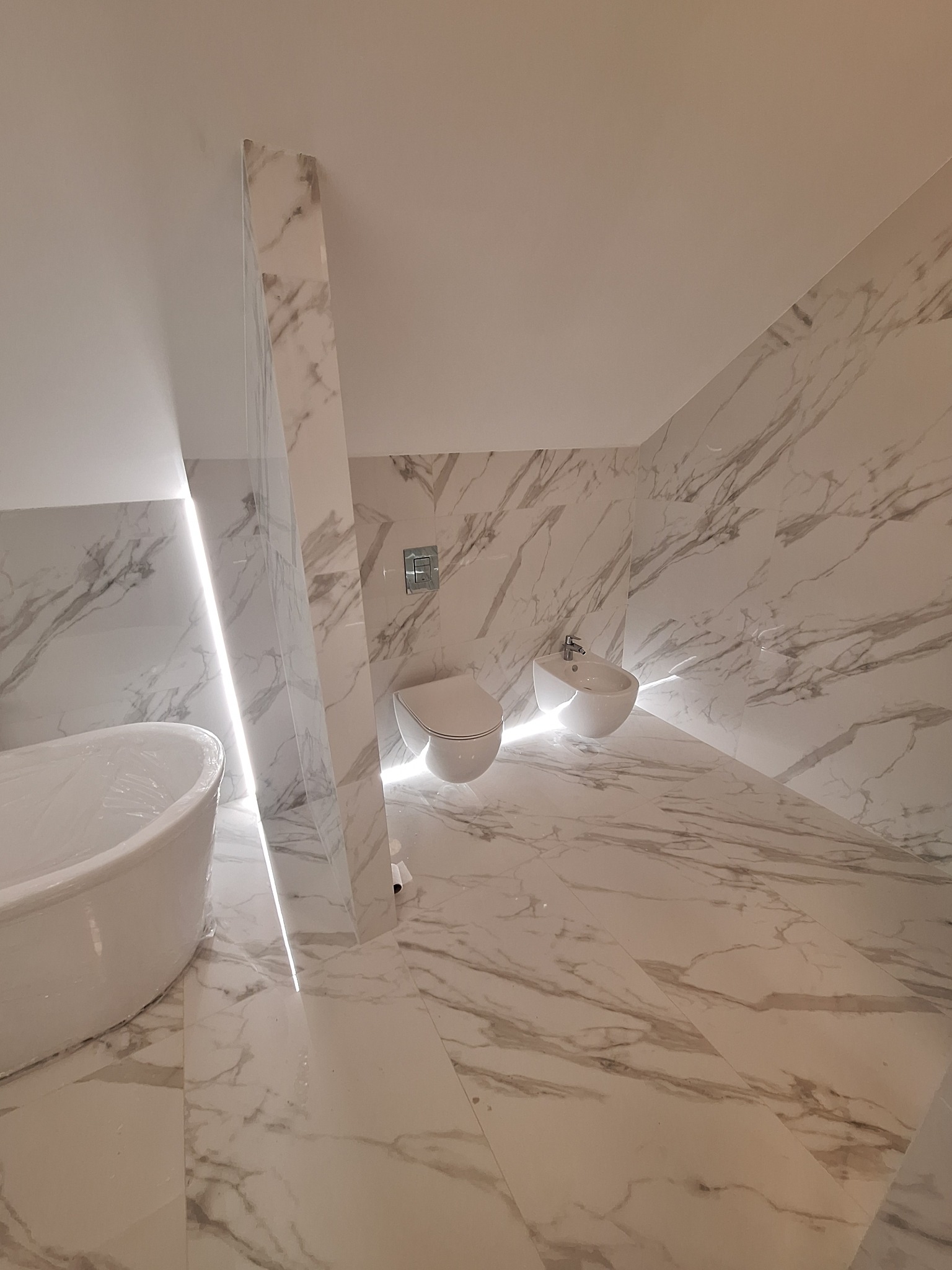 Wykończona łazienka z marmurowymi płytkami, białą wanną wolnostojącą, toaletą i bidetem. Oświetlenie LED wzdłuż ściany działowej.