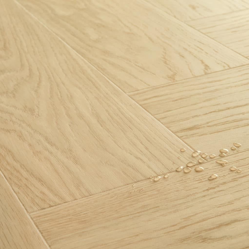 Jasne, drewniane panele ułożone we wzór jodełki francuskiej, z kroplami wody na powierzchni, ukazujące wodoodporność podłogi.